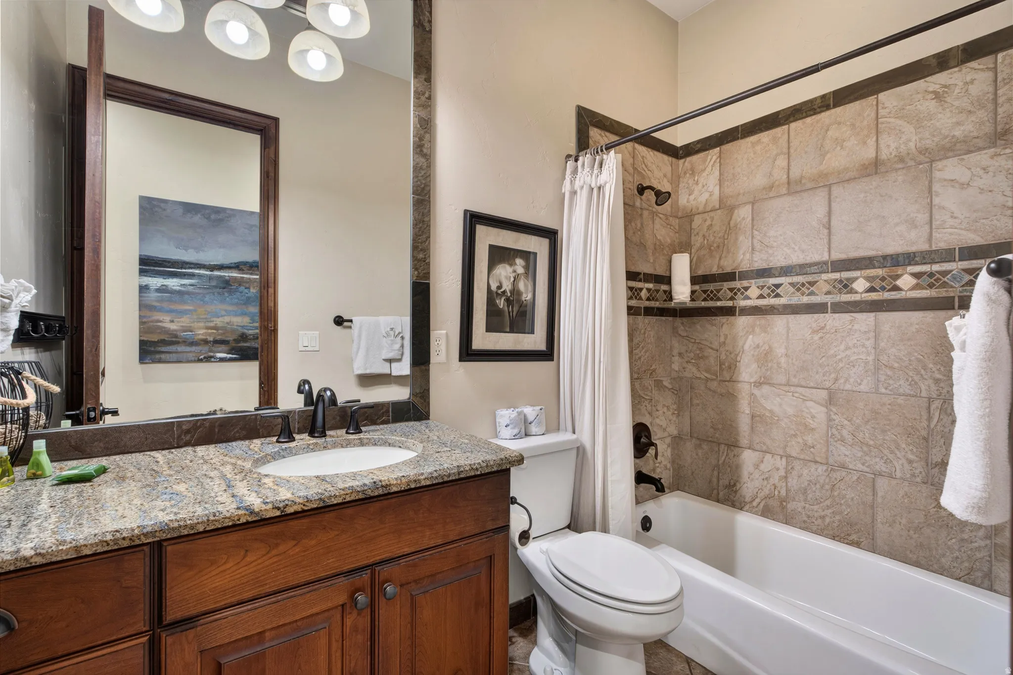 Stonework tile shower surround with an accent band, bathtub, vanity with a granite countertop, undermount sink, and wood-finish cabinetry