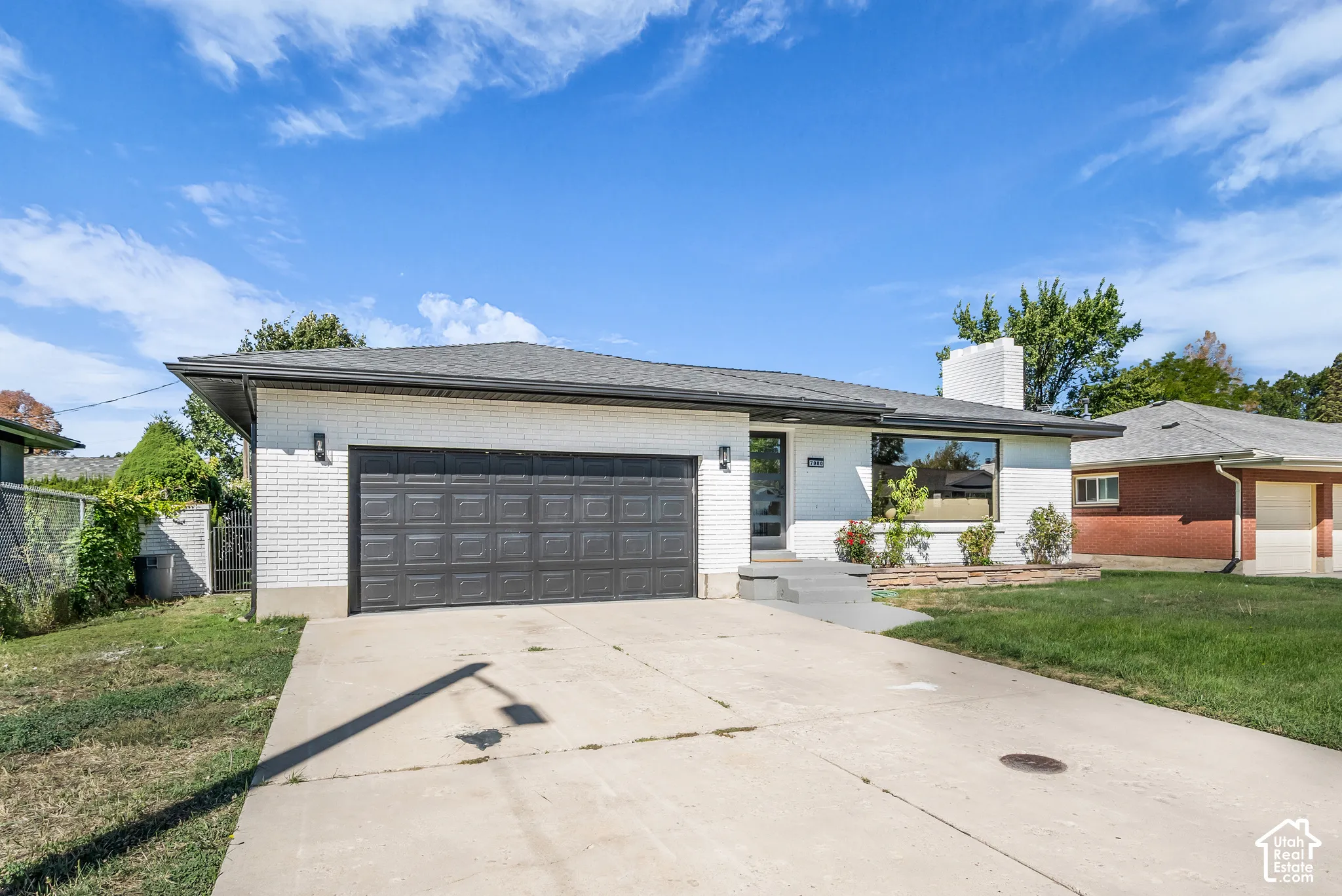 White brick exterior, dark grey shingle roof, two-car garage with a panel door, large front window, and concrete driveway