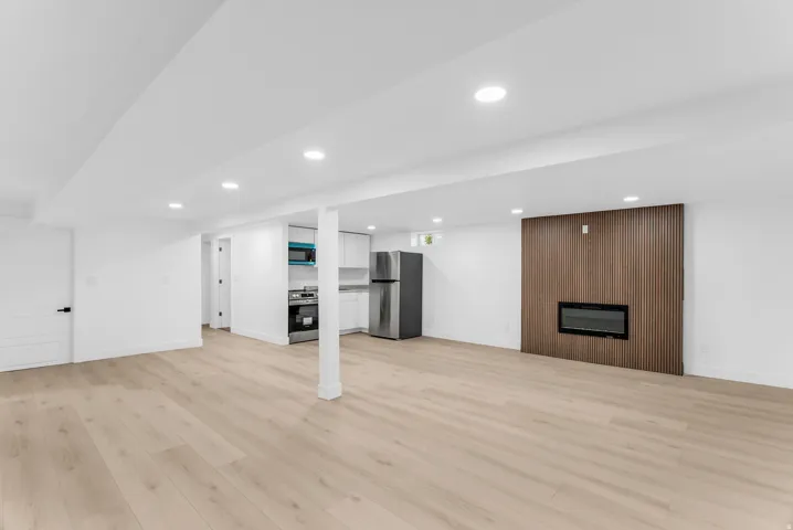 Spacious basement family room featuring wood-finish flooring, recessed lighting, a modern electric fireplace with a slatted wood accent wall, and a compact kitchen area with stainless steel appliances