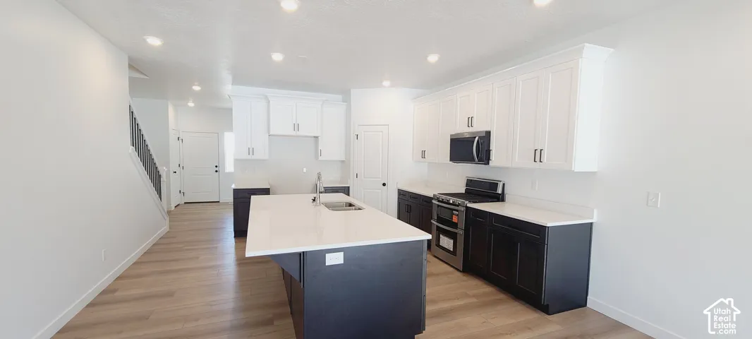 Contemporary kitchen featuring an expansive center island, stainless steel appliances, white upper cabinetry, dark lower cabinetry, and wood-finish flooring