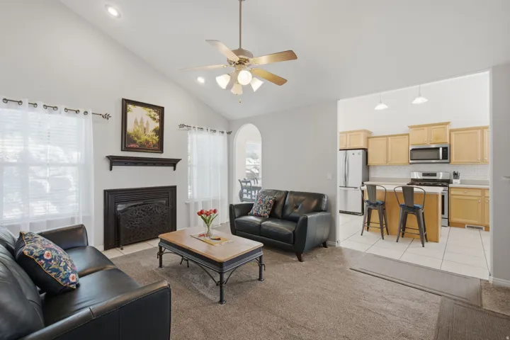 Vaulted ceiling with recessed lighting and ceiling fan, living area with fireplace and dark wood mantel, and kitchen with light wood cabinetry and stainless steel appliances