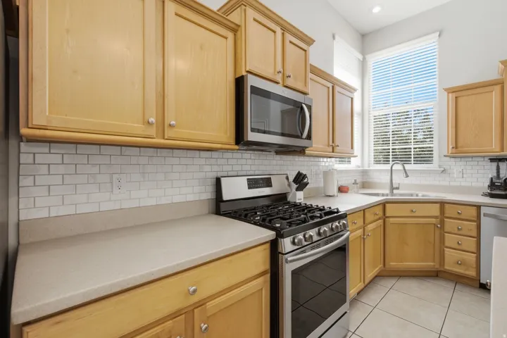 Wood-finish cabinetry, stainless steel appliances, subway tile backsplash, light-toned countertops, and ceramic tile flooring