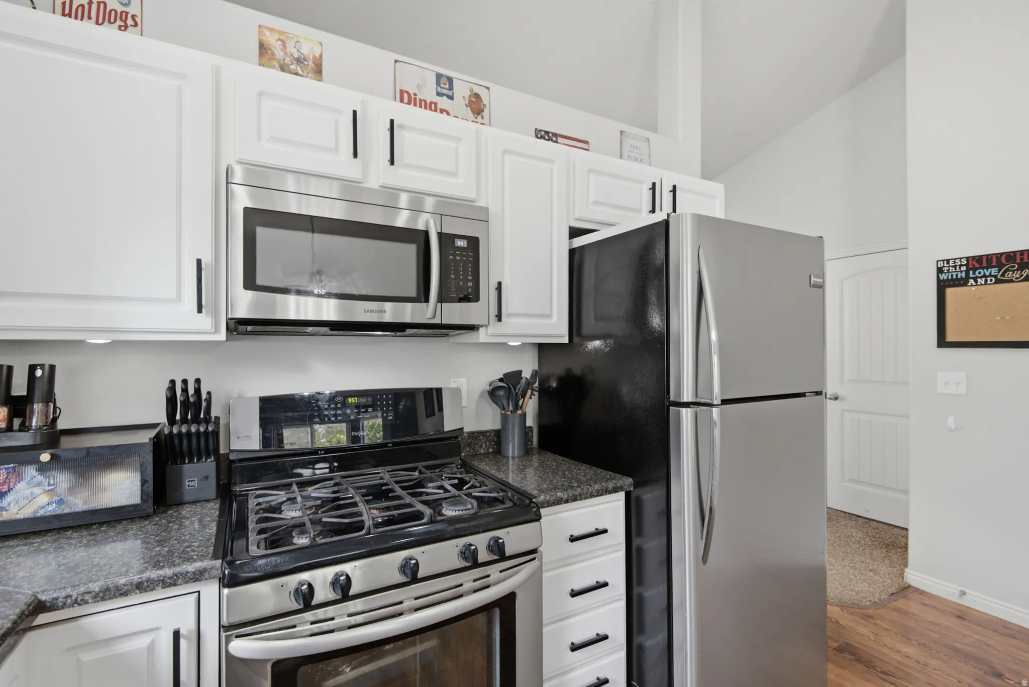 White cabinetry with black hardware, stainless steel appliances, dark granite countertops, and a vaulted ceiling