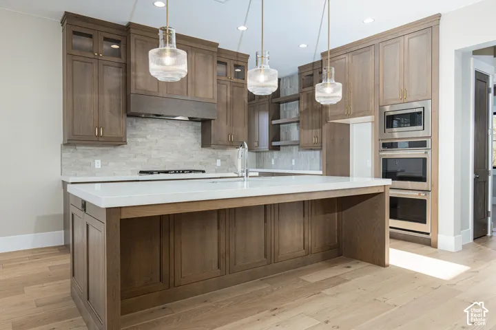 Chef's kitchen featuring custom wood cabinetry with glass-front upper cabinets, a large center island with white quartz countertop, stainless steel gas cooktop, dual wall-mounted ovens, and pendant lighting