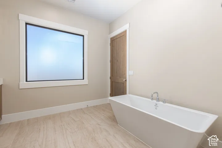 Freestanding soaking tub with chrome fixtures, large window with white trim, wood-finish door, and tile flooring