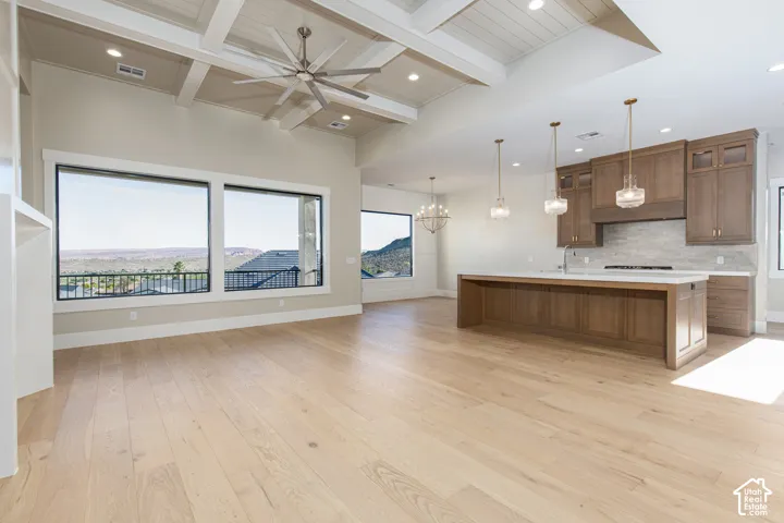 Open-concept living and kitchen area featuring light wood-finish flooring, coffered ceiling with ceiling fan, stainless steel appliances, wood cabinetry with island, pendant lighting, and expansive windows with scenic backdrop