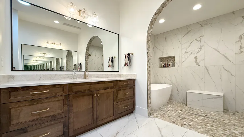 Wood-finish vanity with a light-toned countertop, large rectangular mirror, freestanding soaking tub, and a walk-in shower with pebble tile flooring and a built-in bench