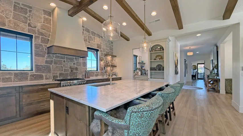 Expansive kitchen island with a light-toned countertop, exposed wood ceiling beams, stone accent wall, wood-finish flooring, and recessed lighting