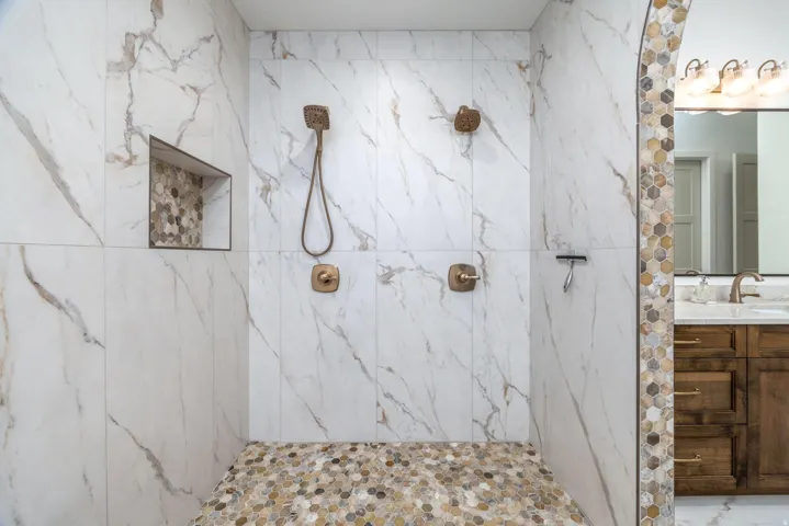Shower with large format marble-look tile walls, pebble stone flooring, built-in niche with hexagonal tile, and dual bronze shower fixtures