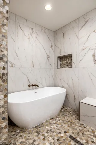 Freestanding soaking tub with a wall-mounted bronze faucet, large format marble-look tile walls, hexagonal pebble tile flooring, and a recessed shower niche with accent tile