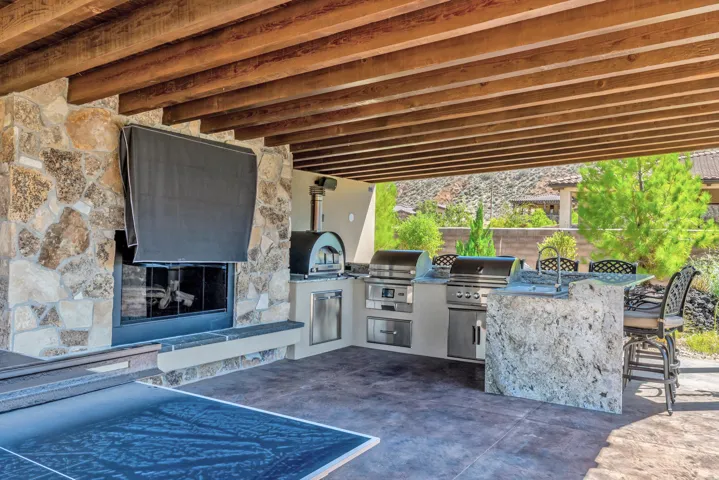 Covered outdoor patio featuring exposed wood beams, a stone-clad fireplace, and a built-in outdoor kitchen with a grill, and sink
