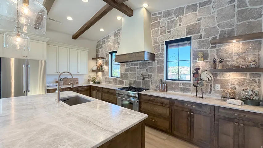 Vaulted ceiling with exposed wood beams, stainless steel appliances, stone accent wall, custom range hood, and an oversized kitchen island with an undermount sink