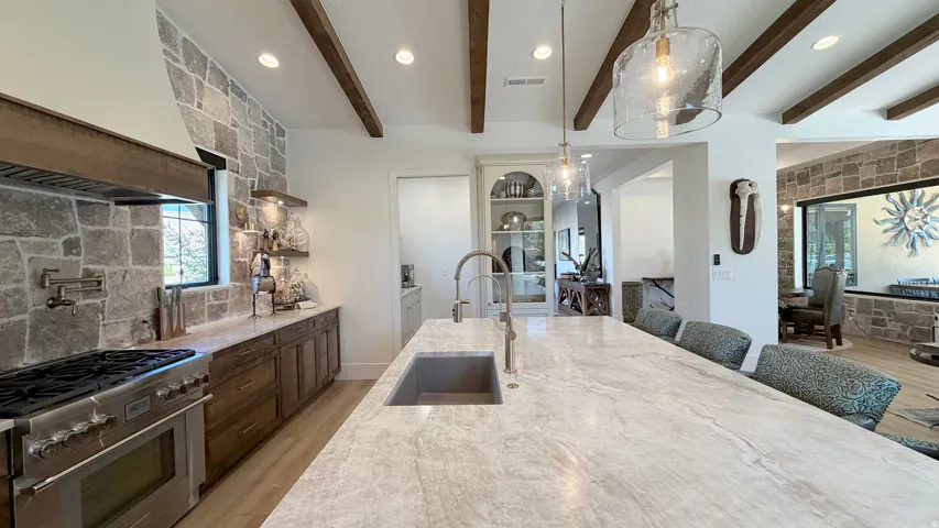 Gourmet kitchen featuring a large stone accent wall, wood-beam ceiling, spacious island with integrated sink, stainless steel professional-grade range, and built-in wood cabinetry