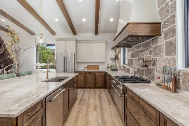 Vaulted ceiling with exposed wood beams, stone accent wall, large island with sink, stainless steel appliances, and wood-finish flooring