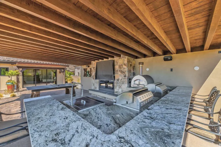 Covered outdoor patio featuring exposed wood beams, a stone-clad fireplace, and a built-in outdoor kitchen with a pizza oven, grill, and sink