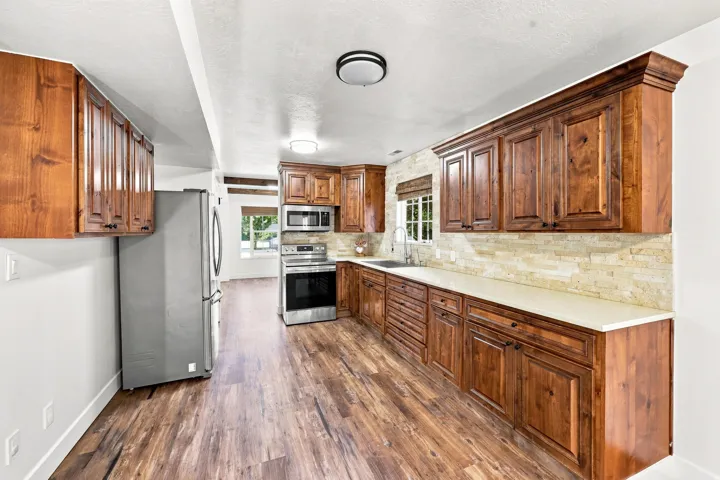 Wood-finish flooring, wood cabinetry with crown molding, light-toned stone backsplash, white countertops, and stainless steel appliances