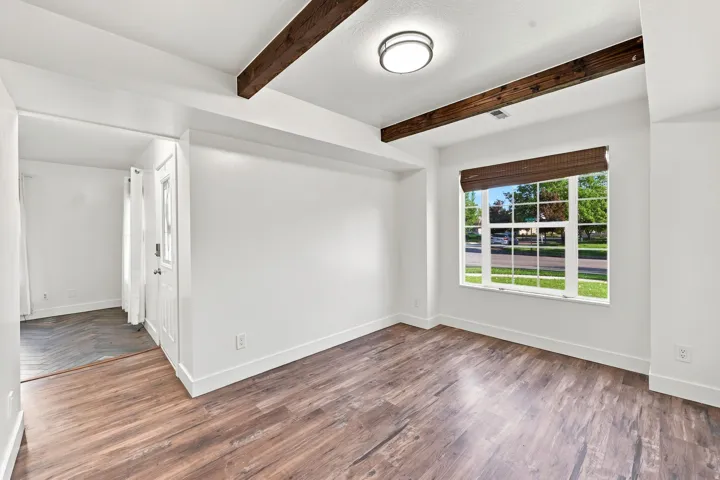 Spacious room featuring wood-finish flooring, exposed ceiling beams, and a large window with a woven shade