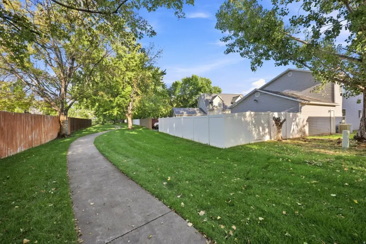 Paved pathway alongside a manicured lawn, bordered by a privacy fence and mature trees