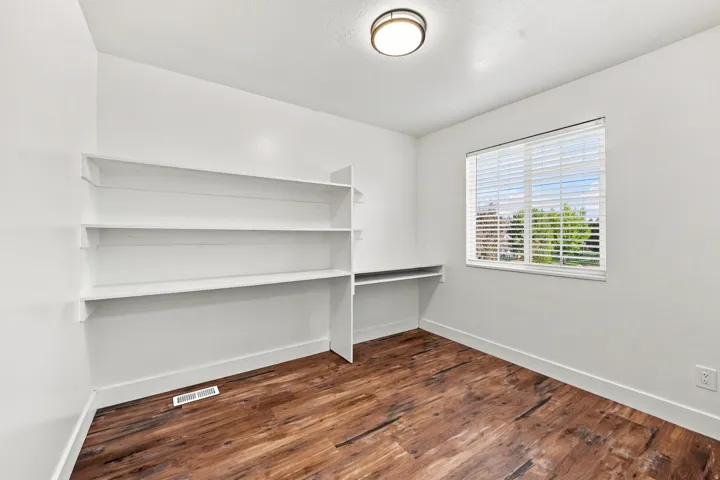 Built-in shelving, wood-finish flooring, recessed desk area, window with blinds, and flush-mount ceiling light