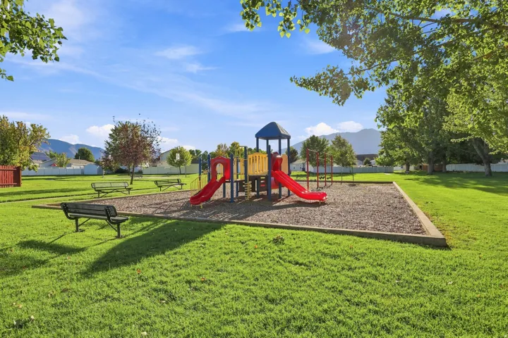 Expansive green space featuring a playground with a slide and climbing structure, multiple park benches, and mature trees