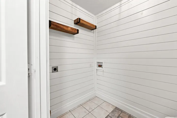 White shiplap walls with two wooden floating shelves, tile flooring, and utility hookups