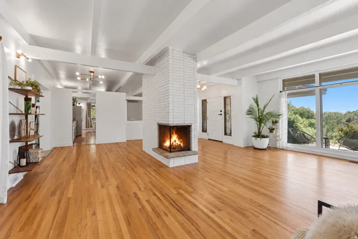 Wood-finish flooring throughout, prominent exposed beam ceiling, white brick fireplace with a glass enclosure, built-in open shelving, and expansive windows