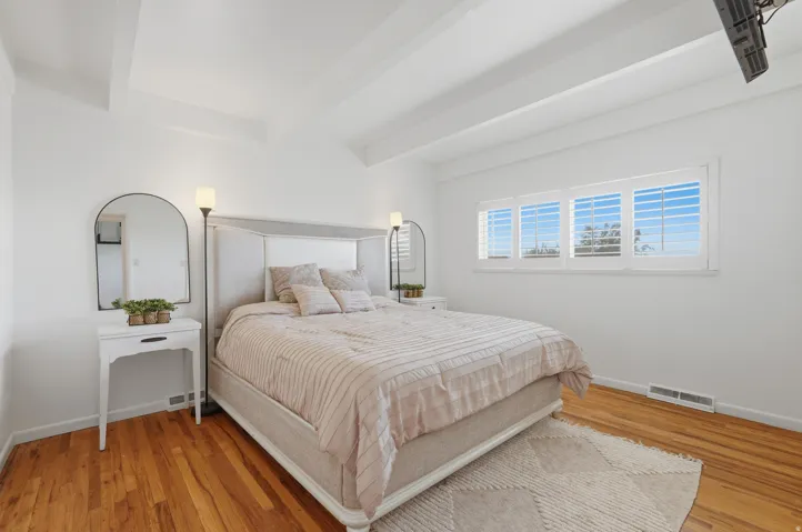 Primary bedroom. Hardwood flooring, white walls, exposed ceiling beams, multiple windows with white shutters