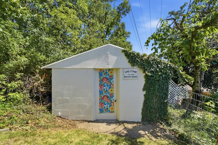 Block construction outbuilding with a peaked roof, featuring a decorative door and established perimeter greenery