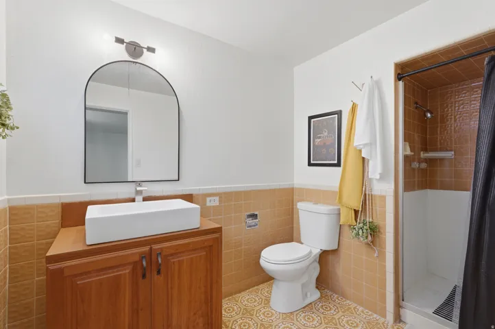 Bathroom #3.  Rectangular vessel sink with a single-handle faucet, wood-finish vanity with cabinet storage, arched mirror, tiled shower enclosure, and pattern-tiled flooring