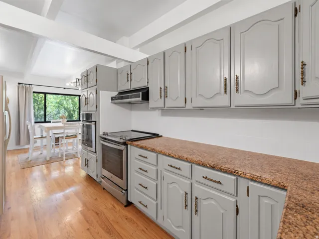 Wood-finish flooring, exposed ceiling beams, light gray cabinetry, built-in double oven, and a large window