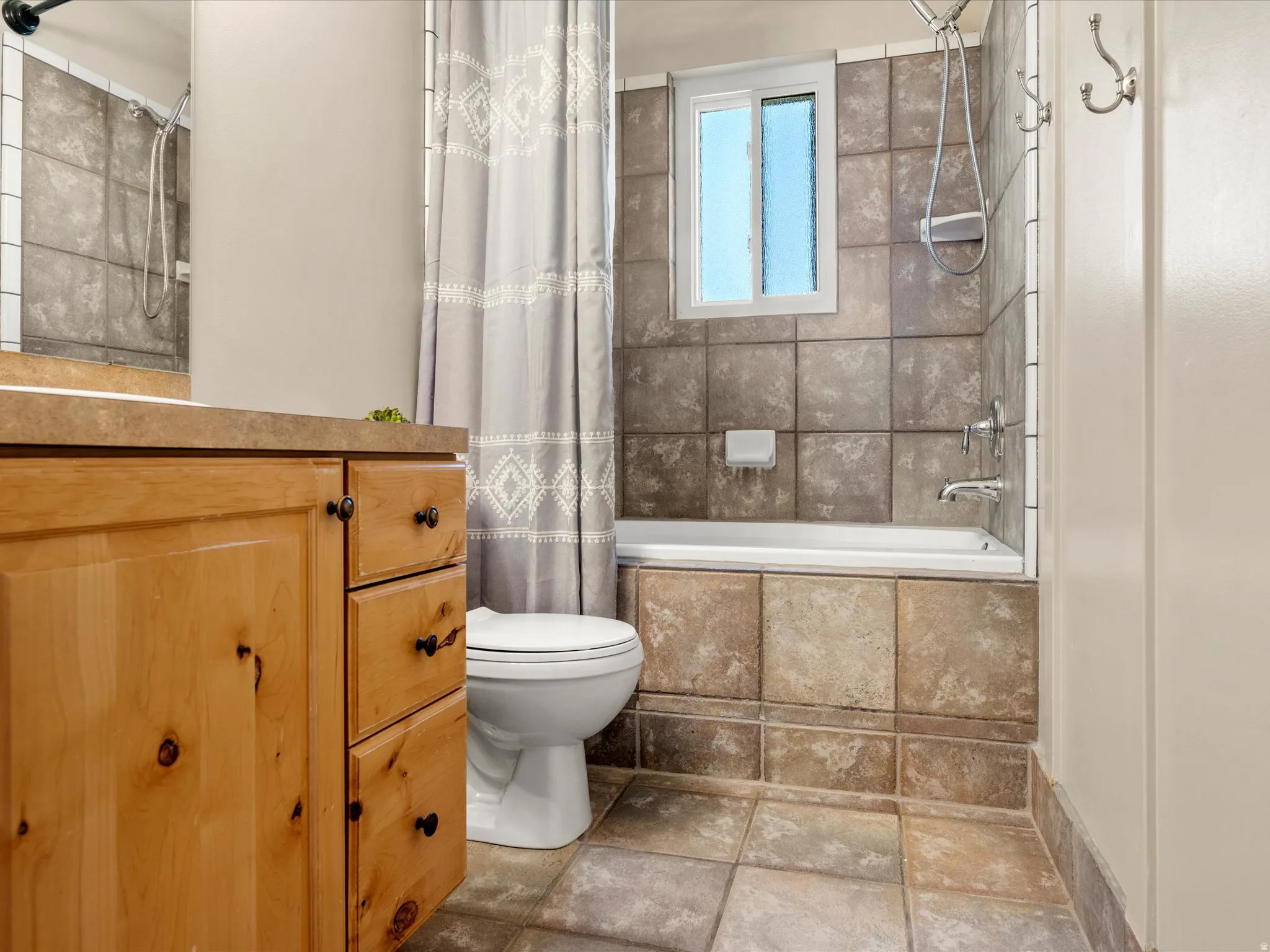 Wood-finish vanity with countertop, tiled flooring, tiled bath and shower surround, and a white framed window