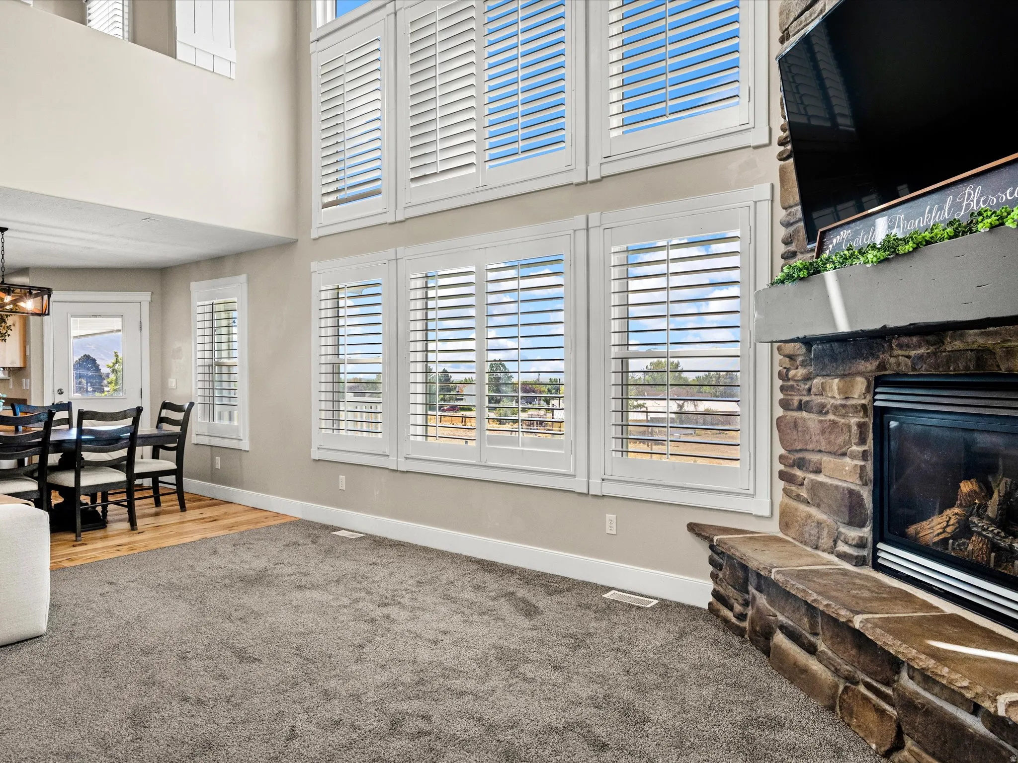 Floor-to-ceiling windows with white plantation shutters, stone-clad fireplace with hearth seating, wood-finish flooring, and recessed lighting