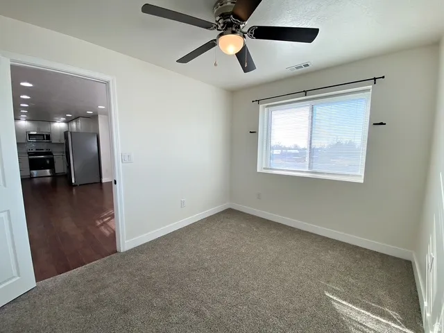 Neutral tone wall paint, flush mount ceiling fan with light, window with blinds, carpeted flooring, and wood-finish flooring visible through doorway