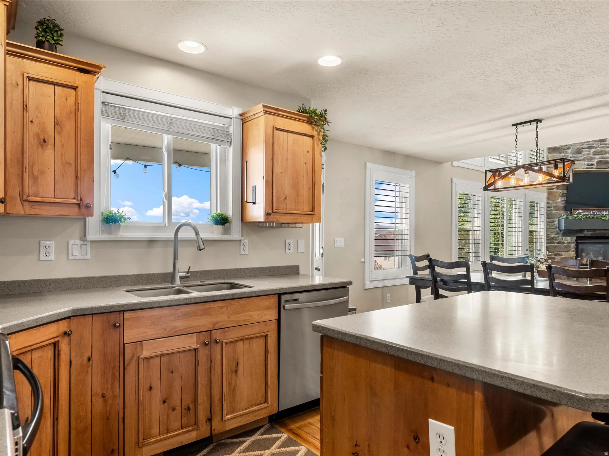 Wood cabinetry with integrated handles, stainless steel dishwasher, double basin sink with gooseneck faucet, light-toned countertops, and recessed lighting