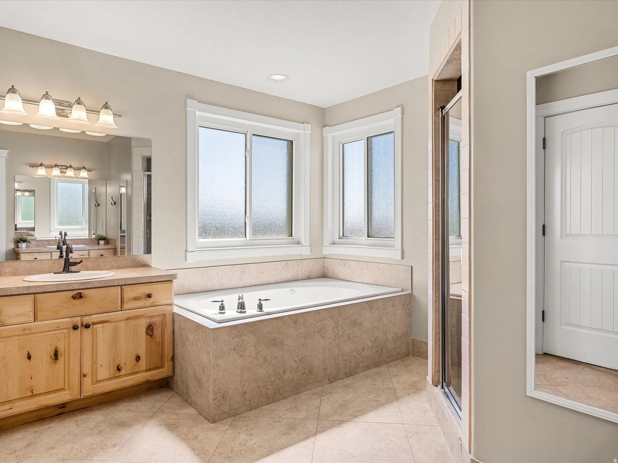 Bathroom featuring a built-in soaking tub, tiled surround, dual windows, wood-finish vanity with countertop, and a glass-enclosed shower