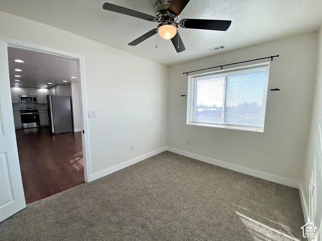 Neutral tone wall paint, flush mount ceiling fan with light, window with blinds, carpeted flooring, and wood-finish flooring visible through doorway