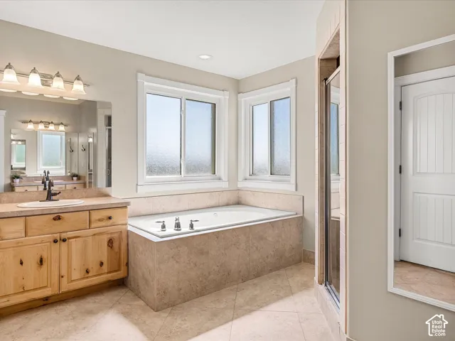 Bathroom featuring a built-in soaking tub, tiled surround, dual windows, wood-finish vanity with countertop, and a glass-enclosed shower