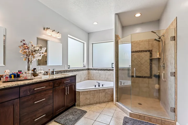 Dual vanity with stone countertop, built-in corner soaking tub, glass-enclosed shower, and tile flooring