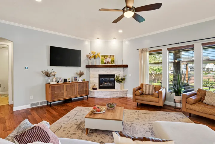 Hardwood flooring, corner stone fireplace with wood mantel, large windows with blinds, recessed lighting, and a ceiling fan
