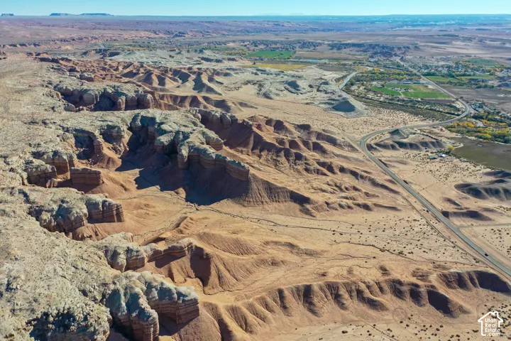 Expansive desert landscape featuring layered rock formations, a winding highway, and distant green areas