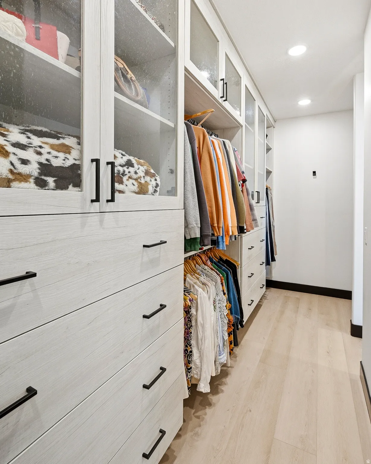 Custom closet system featuring wood-finish cabinetry with glass-front uppers, matte black hardware, integrated shelving, and recessed lighting
