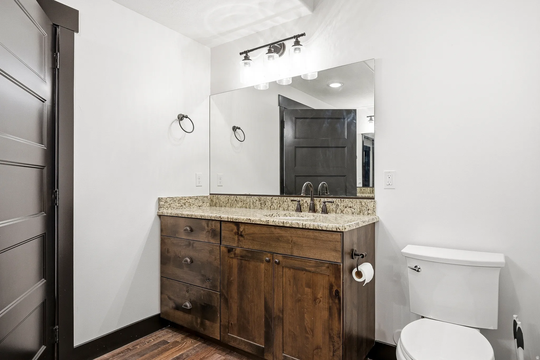 Wood-finish flooring, dark wood vanity with ample storage, light-colored stone countertop, dual-handle faucet, and an overhead vanity light fixture