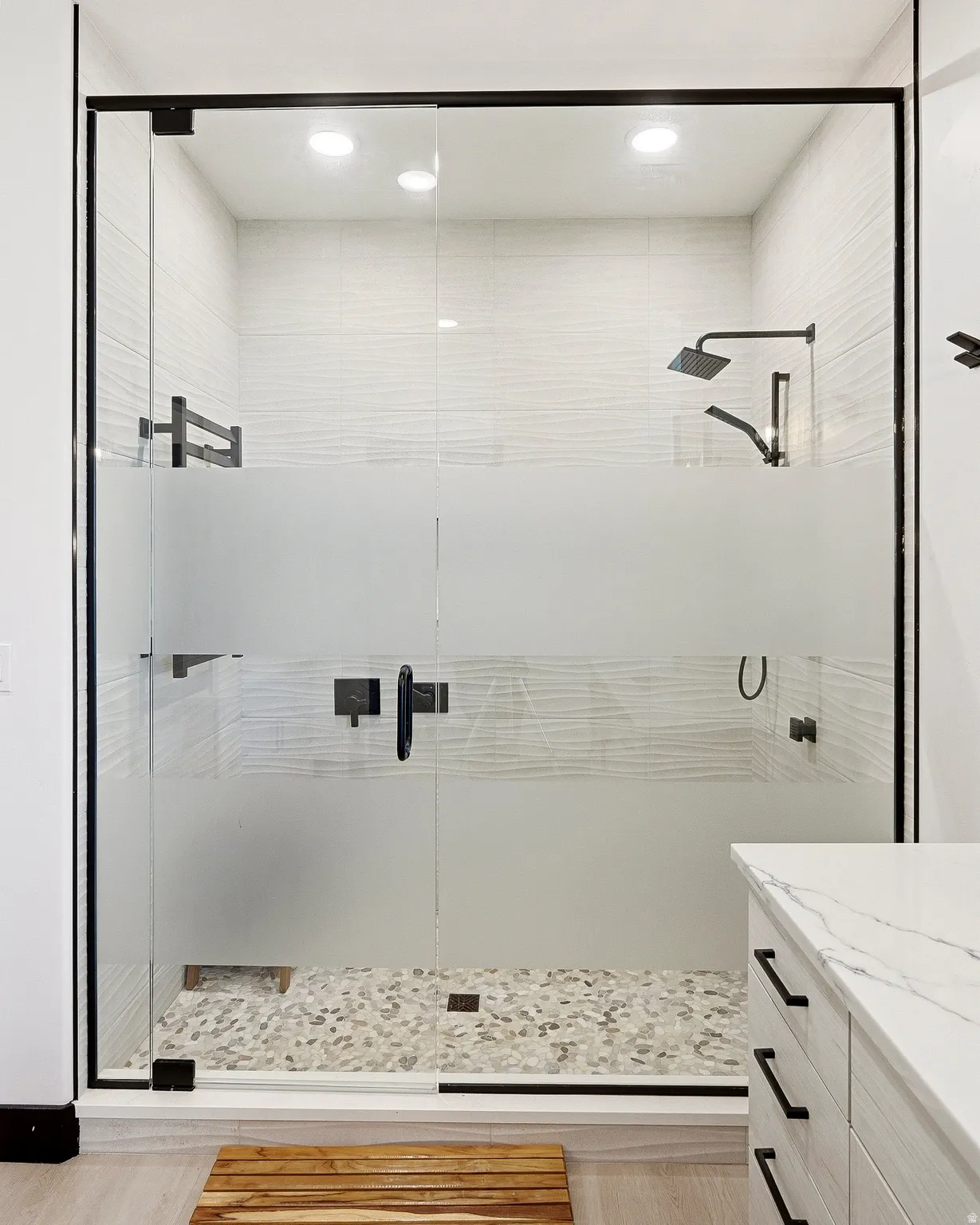 Frosted glass shower enclosure with matte black fixtures, textured tile walls, pebble flooring, and recessed lighting