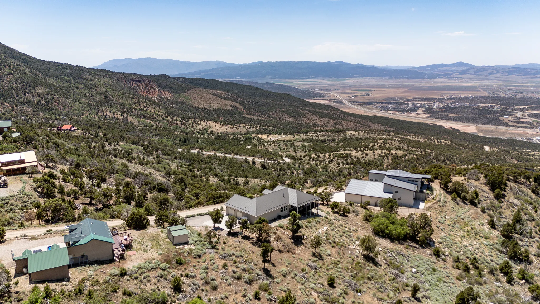 Expansive elevated property featuring a beige exterior, covered porch, and multi-pitched roofline, situated on a sloped hillside with distant mountainous terrain