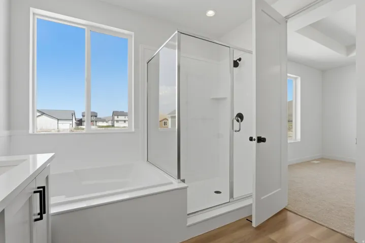 Built-in bathtub with a large window, glass-enclosed shower with black fixtures, white vanity with black hardware, and wood-finish flooring