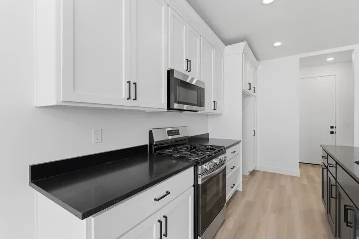 White shaker cabinetry, black countertops, stainless steel gas range, stainless steel microwave, and wood-finish flooring