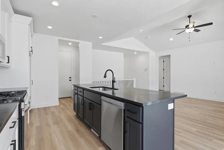 Kitchen island with a dark countertop and integrated sink, stainless steel dishwasher, white upper cabinetry, and wood-finish flooring
