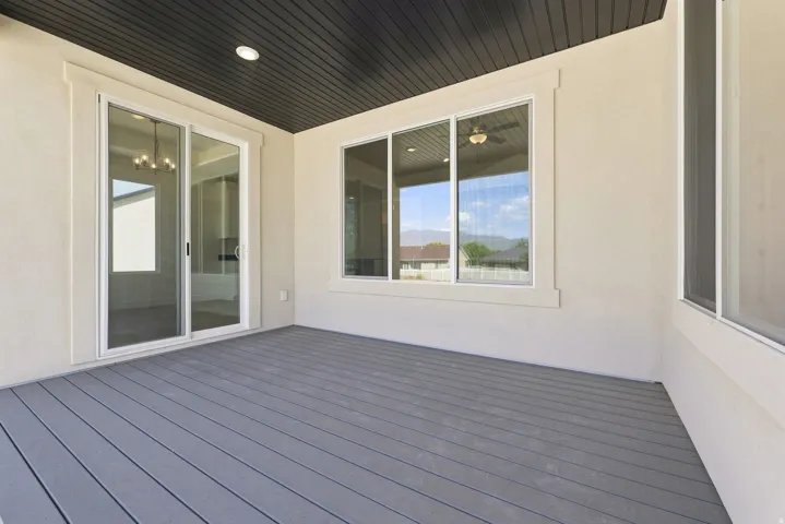 Covered outdoor patio with gray composite decking, white sliding glass door, white framed windows, and a dark soffit ceiling with a recessed light