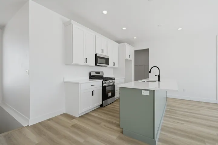Kitchen featuring white shaker cabinetry, stainless steel appliances, a light-toned island with a sink and matte black faucet, and wood-finish flooring