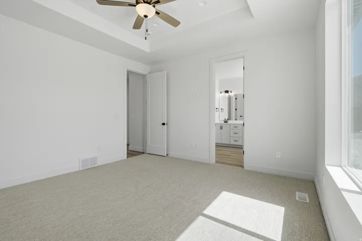 Tray ceiling with a ceiling fan, plush carpeting, large window, and a passage to a bathroom with a dual vanity